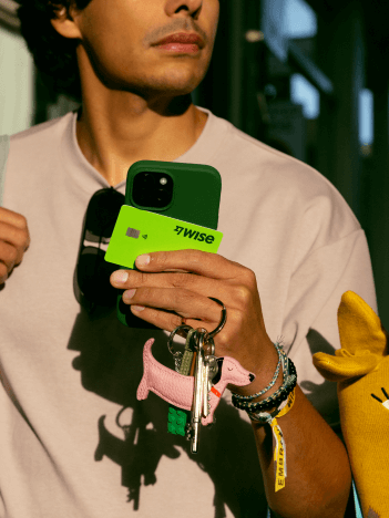 Un homme marche dans une rue commerçante. Il tient dans sa main un téléphone, des clés, des lunettes de soleil et une carte Wise.