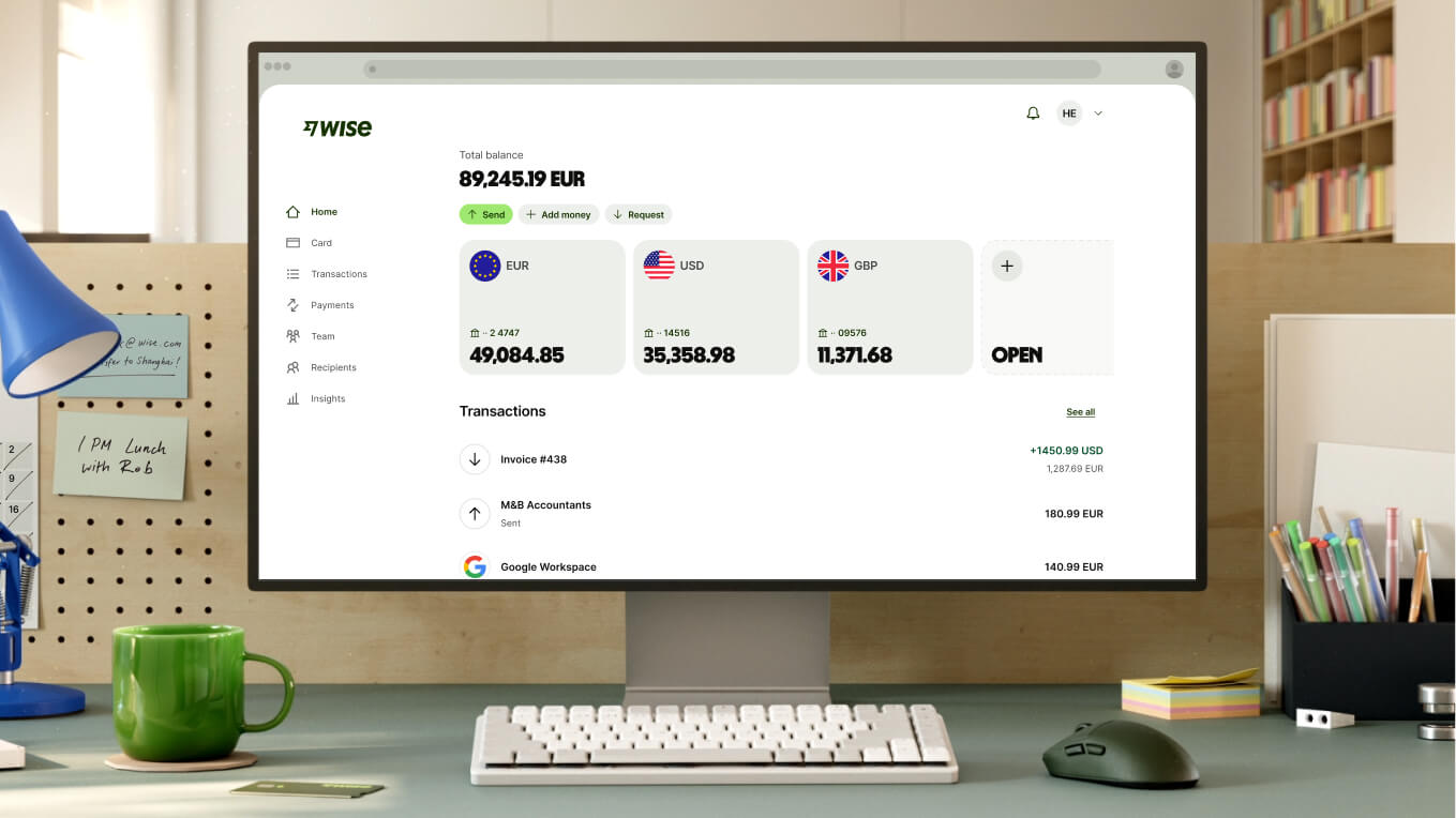 Un gros plan sur un espace de travail moderne montre un écran d'ordinateur blanc affichant le tableau de bord Wise. L'écran indique un solde total de 89 245,19 EUR, réparti entre trois comptes principaux : en EUR (49 084,85), en USD (35 358,98) et en GBP (11 371,68). Une option permettant d'ouvrir un nouveau compte apparaît à droite. Les transactions récentes sont listées en dessous. Devant l'écran se trouvent un clavier blanc compact, une souris gris foncé et une tasse en céramique vert vif.