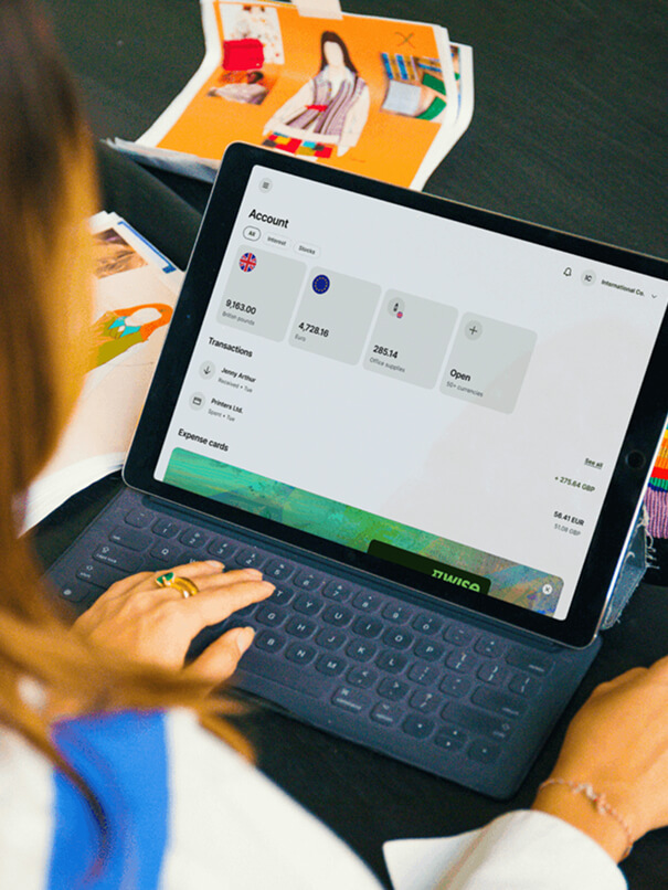 Une personne, vue de dos par-dessus son épaule, est en train de taper sur une tablette. L'écran de la tablette affiche un tableau de bord intitulé « Compte » avec les balances, les transactions et les « Cartes de dépenses ».