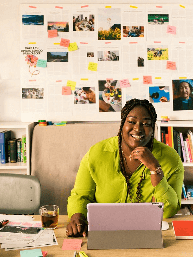 Une femme souriante en chemise verte est assise à un bureau. Devant elle, une tablette violette repose sur un étui gris. En arrière-plan, un mur tapissé de photos, de croquis et de post-it colorés évoque un environnement de travail créatif. Les mots « POUR LES STARTUPS » apparaissent en blanc en haut de l'image, tandis qu'un cercle vert avec un signe « + » blanc est visible en bas à droite.